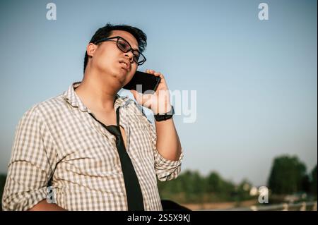 Giovane uomo d'affari asiatico in camicia e cravatta con pantaloncini e occhiali da vista in piedi e utilizza lo smartphone in spazi esterni, fotocopie Foto Stock
