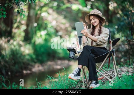 Giovane donna seduta su una sedia e utilizza una videochiamata con un tablet mentre campeggi nel parco naturale, spazio di copia Foto Stock