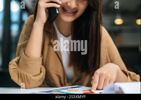 Primo piano, una giovane donna usa lo smartphone mentre lavora su un computer portatile e documenti o presenta un grafico di crescita del progetto al bar Foto Stock
