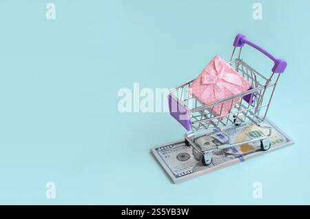 La confezione regalo di un piccolo carrello si trova su una bolletta di un dollaro su sfondo azzurro. La confezione regalo in un piccolo carrello si trova su un dollaro di banconote sulla luce Foto Stock