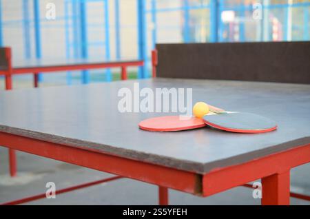 Racchette e palla da vicino sul tavolo da ping pong in un cortile sportivo all'aperto. Concetto di sport attivi e allenamento fisico. Racchette e palla sul tavolo da ping pong Foto Stock