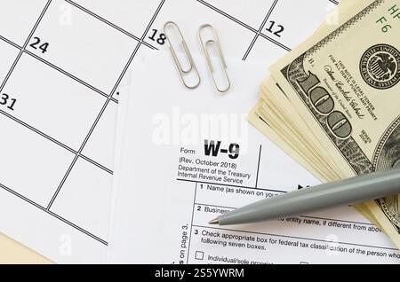 Modulo IRS W-9 richiesta di numero di identificazione contribuente e certificato vuoto si trova con la penna e molte banconote da cento dollari sulla pagina del calendario. Tassa Foto Stock