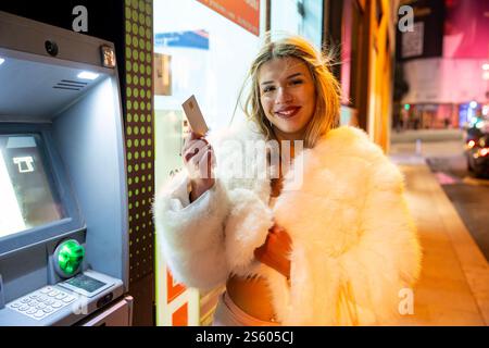 Giovane donna alla moda che ritira soldi da un bancomat di notte in città Foto Stock