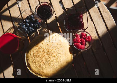 Deliziosi pancake serviti su un rustico tavolo in legno abbinati a lamponi freschi, mirtilli e varie gustose marmellate. Perfetto per una colazione accogliente o. Foto Stock