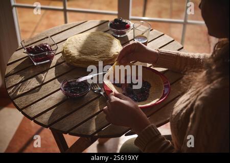 Un ambiente accogliente per la colazione, dove una persona prepara crepes con marmellata e frutti di bosco freschi, catturati dalla luce naturale, evocano un'atmosfera calda e invitante. Foto Stock