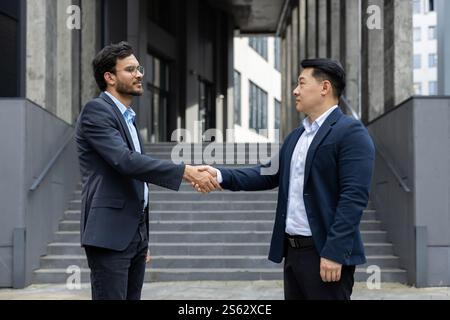 Due uomini seri in abiti da lavoro si salutano stringendo la mano fuori da un edificio di uffici. Persone d'affari diverse che camminano per la città. Foto Stock