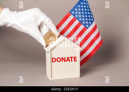 Mano che mette la moneta Crypto Bitcoin nella casa del salvadanaio per donare su American Flag. Costruisci una casa con Crypto. Donazione cripto per la California Foto Stock