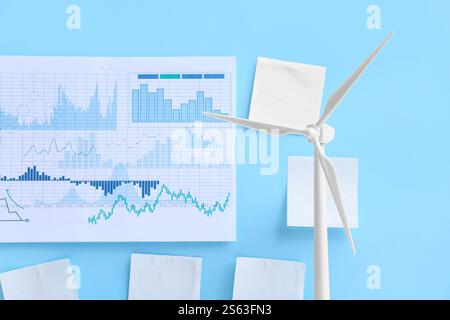 Modello di turbina eolica con diagrammi e note adesive vuote su sfondo blu, primo piano. Concetto di energia verde Foto Stock