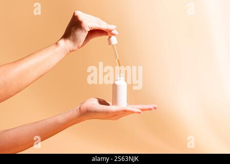 Le mani femminili tengono in mano il flacone contagocce per siero bianco su sfondo arancione beige con luce solare, spazio di copia. Olio essenziale, siero retinolo facciale. cosmetici per la cura della pelle Foto Stock