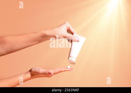 Le mani delle donne tengono e applicano crema idratante cosmetica per la cura della pelle su sfondo arancione beige con la luce del sole. Tubo bianco, marchio cosmetico del prodotto Foto Stock