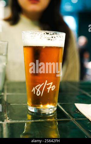 Barcellona, Spagna - 10 gennaio 2025; primo piano di un bicchiere di birra ghiacciata su un tavolo piastrellato verde con una giovane donna sullo sfondo Foto Stock
