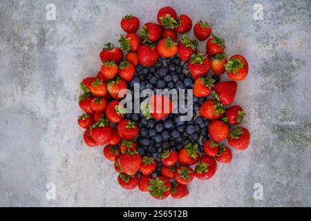 Vista dall'alto delle fragole e dei mirtilli maturi, splendidamente disposti in un design circolare su sfondo grigio testurizzato Foto Stock