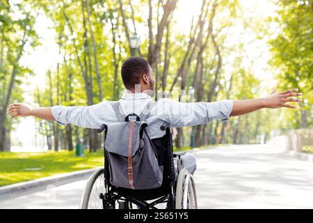 Vista posteriore di un giovane uomo nero disabile in sedia a rotelle che si allarga le braccia, sentendosi felice e libero a piedi al parco della città Foto Stock