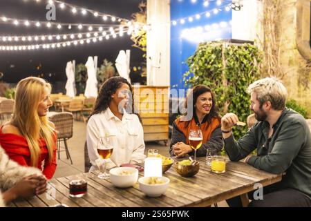 Amici multietnici maschi e femmine che parlano rilassato bevendo alcolici e mangiando spuntini in una terrazza bar serale Foto Stock
