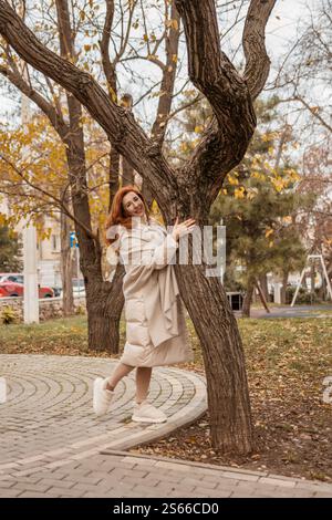 Woman Tree Autumn Park: Elegante rossa posa giocosamente vicino all'albero nel parco autunnale. Foto Stock