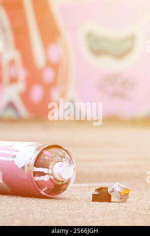 Bomboletta spray spray usata con vernice rosa e bianca e cappucci per spruzzare la vernice sotto pressione si trova sull'asfalto sporco vicino alla parete verniciata nella Foto Stock