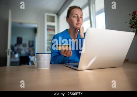 Una donna incentrata sul laptop utilizza la carta di credito per le transazioni online, persa in spese pesanti. Debito di credito Foto Stock