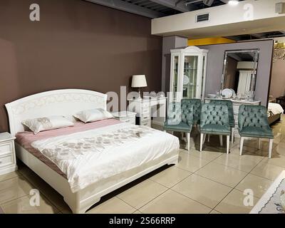 Un'accogliente camera da letto con letto bianco, sedie verdi decorative, armadietto in vetro e illuminazione soffusa che creano un'atmosfera invitante. Foto Stock