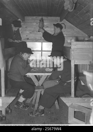 Durante la seconda guerra mondiale. La guerra rendeva difficile importare carbone per riscaldare case e appartamenti. Il legno doveva essere usato al posto del carbone. Nelle grandi città come Stoccolma, si potevano vedere grandi cumuli di legno lungo le strade. A molti uomini fu dato un lavoro temporaneo per lavorare sulla triturazione della legna. Qui ci sono due giovani che lavorano nelle foreste fuori dalla città di Västmanland, Skinnskatteberg. Si stanno prendendo una pausa molto necessaria nelle loro semplici caserme dove non c'è elettricità. Si siedono con i loro cappotti per tenersi al caldo e tre dei giovani fumano tubi. Una lanterna a cherosene è appesa Foto Stock