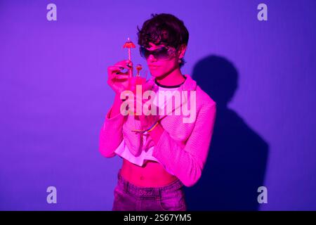 Un giovane elegante regala un cocktail con un piccolo ombrello in una luce al neon viola Foto Stock