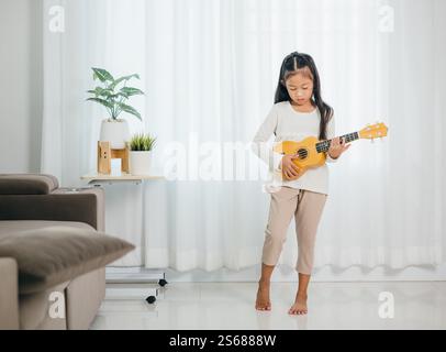 Ritratto di una ragazza asiatica in piedi in un salotto moderno, che suona un ukulele. Il suo brillante sorriso e.. Foto Stock