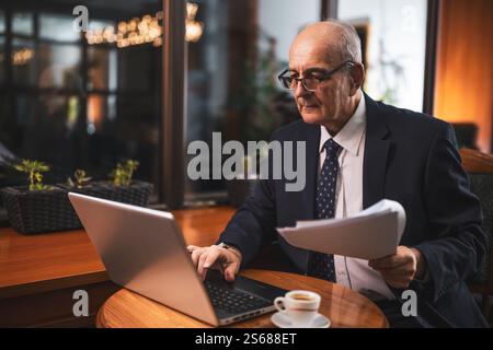 Uomo d'affari anziano concentrato in elegante tuta con occhiali da vista che usano il computer portatile e che esaminano i documenti mentre beve un caffè e si siede al bar. Foto Stock