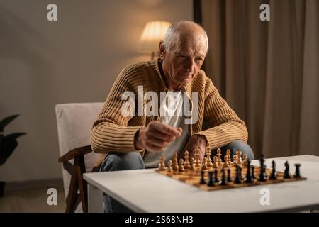 L'uomo anziano ama giocare a scacchi tranquilli a casa, usandolo sia come hobby rilassante che come un modo per rafforzare le sue capacità cognitive. Foto Stock