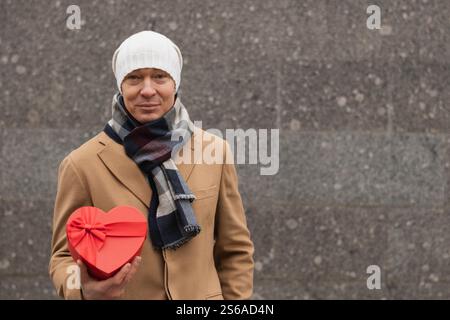 Il bell'uomo felice regala un regalo rosso a forma di cuore con un cappotto beige a San Valentino. Primo piano. Copia spazio. Ritratto all'aperto in città. Foto Stock