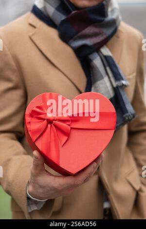 Il bell'uomo felice regala un regalo rosso a forma di cuore con un cappotto beige a San Valentino. Primo piano. Foto Stock