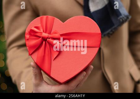 L'uomo felice regala un regalo rosso a forma di cuore con un cappotto beige a San Valentino. Primo piano. Concetto d'amore per il compleanno o l'anniversario. Foto Stock