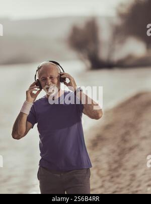 Uomo anziano che si riscalda per fare jogging sulla spiaggia e indossa le cuffie per motivare la musica per l'esercizio fisico. Concetto di stile di vita sano Foto Stock