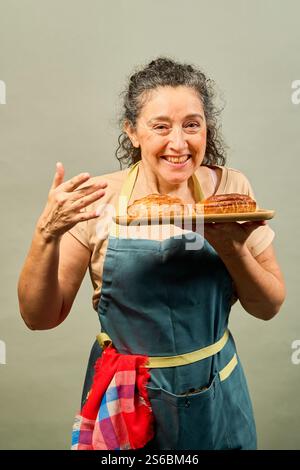 Una donna sta tenendo due dolci su un vassoio di legno e sorride. Indossa un grembiule blu e una sciarpa rossa e bianca Foto Stock