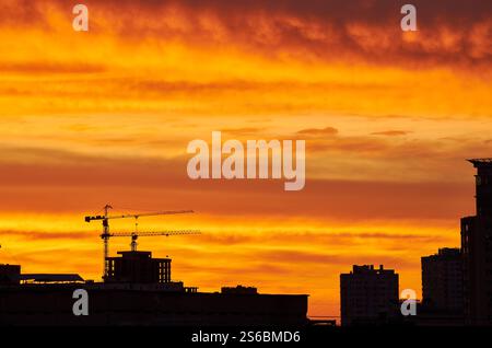 Una gru da costruzione si staglia contro un cielo rosso e arancione intenso. L'orizzonte luminoso crea un contrasto sorprendente con l'elem urbano scuro Foto Stock