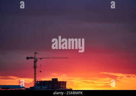 Una gru da costruzione si staglia contro un cielo rosso e arancione intenso. L'orizzonte luminoso crea un contrasto sorprendente con l'elem urbano scuro Foto Stock