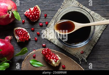 Melassa di melograno in un recipiente di vetro, salsa acida di melograno con frutta fresca matura intera e frazionata su un tavolo rustico in legno (Punica granatum) Foto Stock