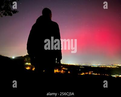 Silhouette di una persona che osserva le rare luci boreali su San Marino, con vivaci sfumature di rosso e rosa che illuminano il cielo notturno e la città sottostante Foto Stock