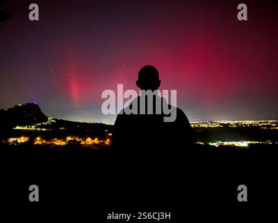 Silhouette di una persona che osserva le rare luci boreali su San Marino, con vivaci sfumature di rosso e rosa che illuminano il cielo notturno e la città sottostante Foto Stock