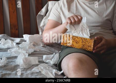 Giovane seduto a letto con tessuti facciali che soffiano il naso Foto Stock