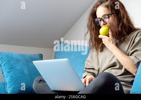 Donna che mangia mela verde mentre usa il suo laptop seduto sul divano di casa. Foto Stock