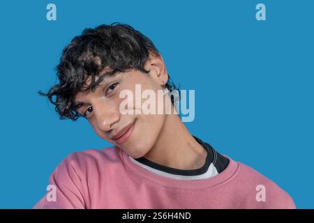 Ritratto di un giovane allegro con capelli ricci e felpa rosa in posa su un vivace sfondo blu Foto Stock
