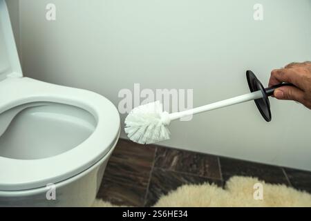 Uno spazzolino bianco con manico nero tenuto vicino a una ciotola bianca, collocato su un pavimento piastrellato con un soffice tappeto beige. Foto Stock