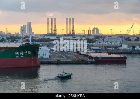 Abu Dhabi, Emirati Arabi Uniti - 3 gennaio 2025: Una vivace scena portuale al tramonto con una grande nave da carico e un piccolo rimorchiatore che navigano nelle acque, struttura industriale Foto Stock