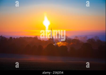 Un villaggio di campagna inglese, circondato dalla nebbia, in una splendida alba autunnale. L'alba dorata trasforma la guglia della chiesa in una silhouette tranquilla. Foto Stock