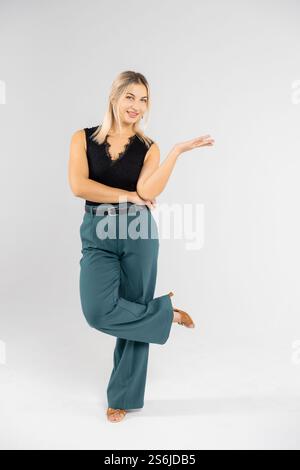 Giovane donna d'affari bionda che mostra qualcosa con la mano su sfondo bianco, con top nero e pantaloni verdi Foto Stock