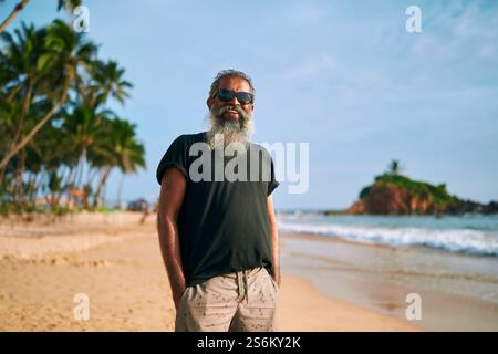L'uomo barbuto anziano ama fare una gita al mare. Indossati con stile con occhiali da sole. Passeggiata rilassante sulle onde dell'oceano. Stile di vita attivo per anziani. Viaggi popolari Foto Stock