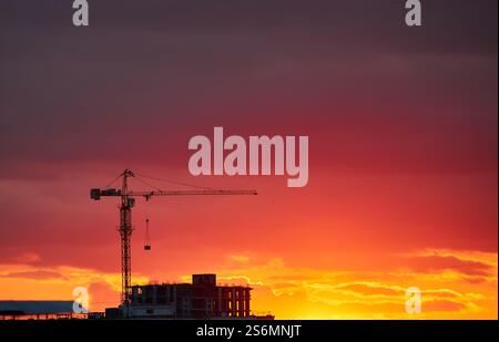 Una gru da costruzione si staglia contro un cielo rosso e arancione intenso. L'orizzonte luminoso crea un contrasto sorprendente con l'elem urbano scuro Foto Stock