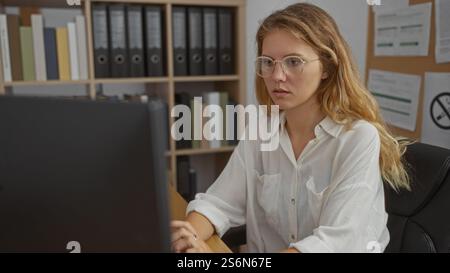 Donna che lavora alla scrivania in ufficio con scaffali e documenti in background, concentrandosi sullo schermo del computer, indossando occhiali da vista e camicia bianca, BL Foto Stock