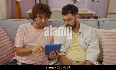 Due uomini seduti insieme su un divano di casa, che si impegnano con un tablet, illustrano il legame con la famiglia e l'interazione tecnologica in un accogliente ambiente del soggiorno Foto Stock