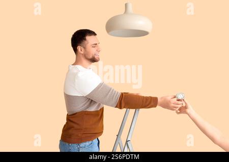 Giovane uomo su scala a cavalletto che prende la lampadina per appendere la lampada da donna su sfondo beige Foto Stock