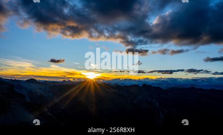 A stunning sunset unfolds over towering mountains, illuminating the sky with vibrant hues of orange and gold. Clouds drift lazily as the sun dips below the horizon, creating a captivating sight. Foto Stock
